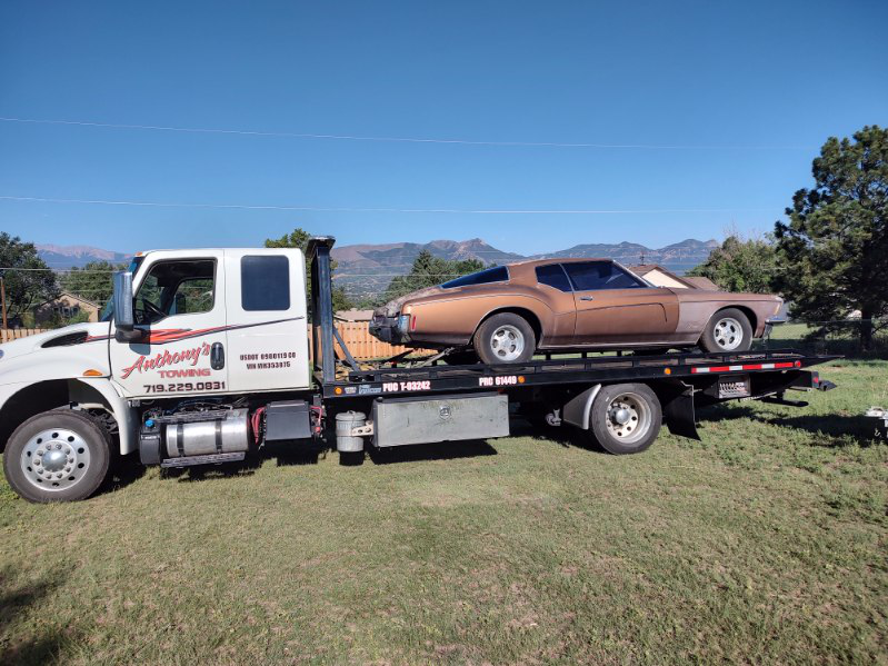 Bronze car on a tow truck placed on a grassy patch with majestic mountains and blue sky in the background.