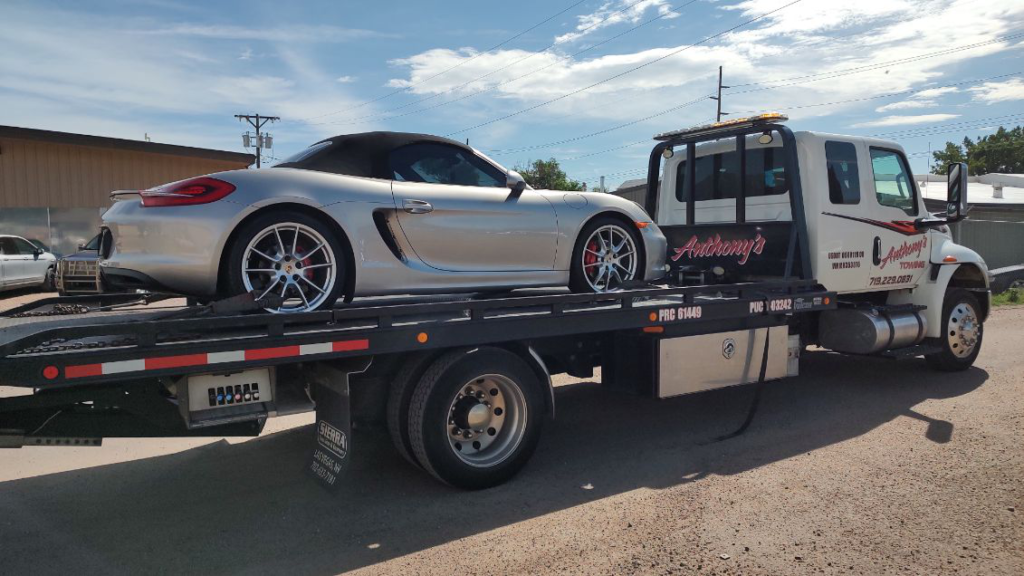 Silver Porsche convertible loaded on a tow truck in a lot, set against a vivid blue sky with scattered clouds.