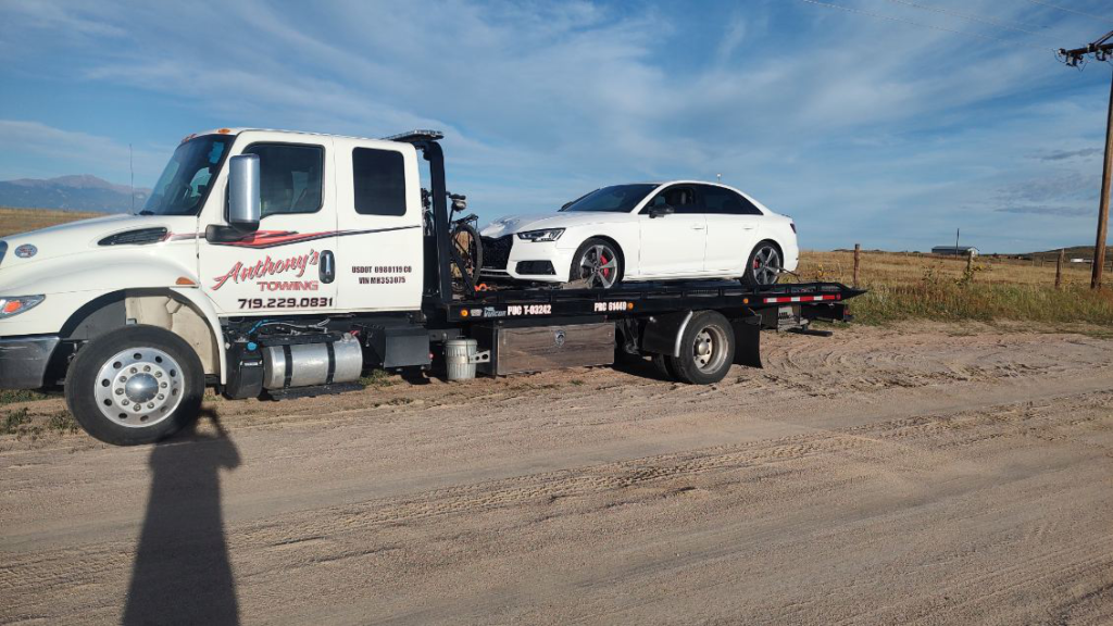 A white sedan is being towed on a tow truck with a sandy landscape, open field, and blue sky in the background.