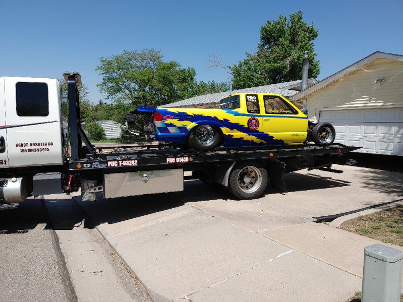 A blue and yellow eclectic pickup truck missing some parts on a tow truck in front of a house.