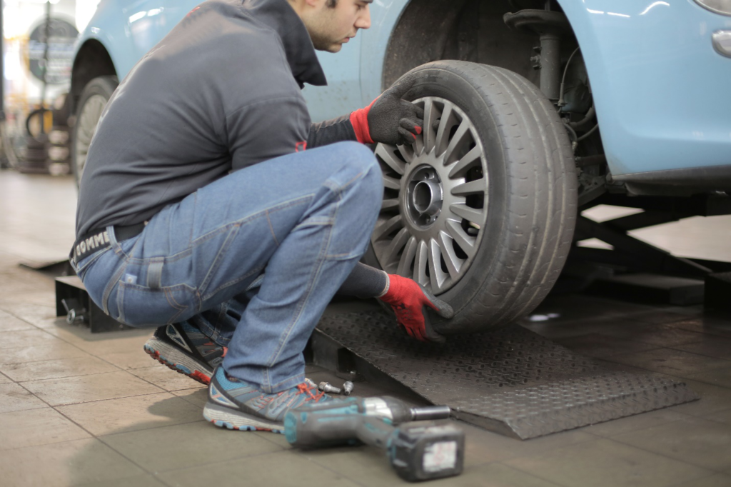A mechanic removes a tire from a light blue car while wearing gloves as his tools lay nearby