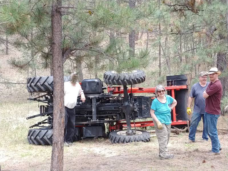 A group of people in a wooded area standing beside an overturned tractor, patiently waiting for Anthony's Towing wrecker services.