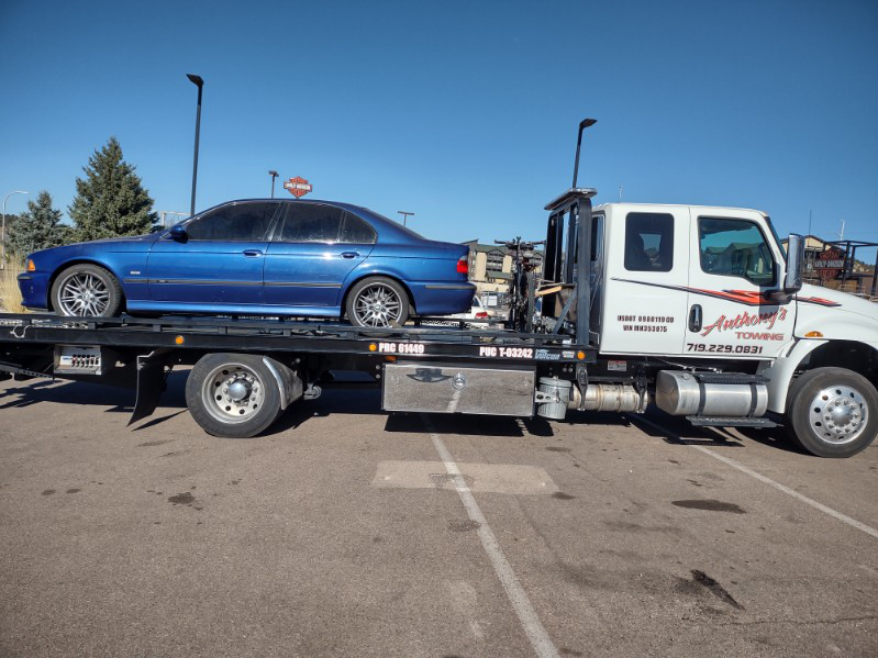 A blue car positioned atop a white Anthony's Towing tow truck in a spacious lot, creating a striking visual contrast.