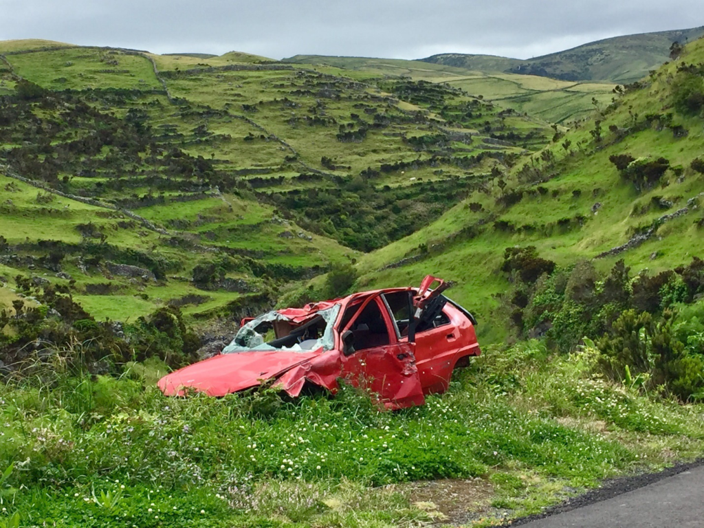 A totaled red car lies in a grassy patch on the side of the road before a descent down a lush green hill.