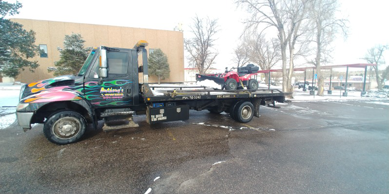 A small red snow removal vehicle with a bulldozer attachment secured on a black Anthony's Towing truck with colorful flame graphics on the front amidst snow on a wet road because everyone needs affordable towing services.