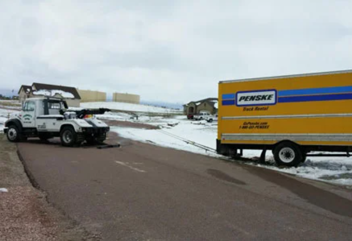 A white tow truck stands on the road while a yellow Penske trailer stands in the snow on the side